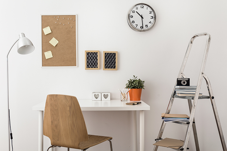 Small office space with wall clock, plant, step ladder and wooden pin board for creative ideas.