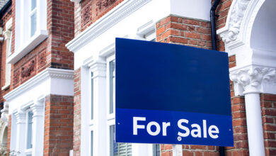 Estate agent 'For Sale' sign board with brick houses in background