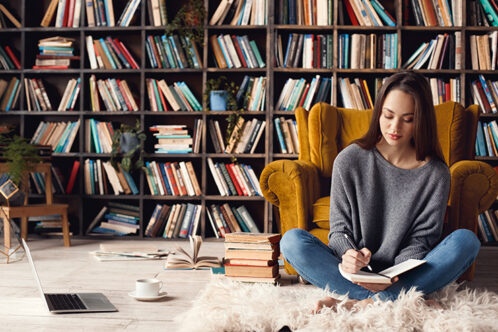 Picture of a woman sitting on the floor writing with a home library in the back
