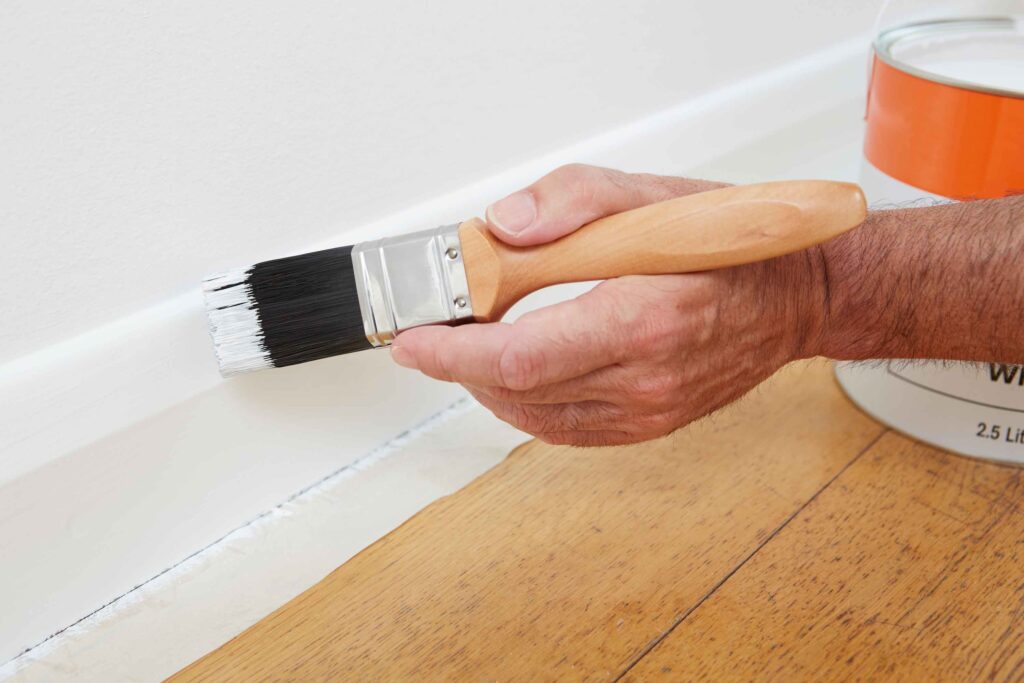 Picture of a hand holding a paintbrush applying white paint to a skirting board
