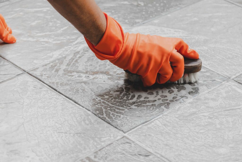 Picture of a hand wearing an orange glove holding a brush scrubbing a floor with soap